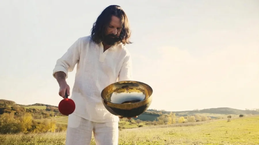 Leo Melcherts in white linen in a hilly meadow, holding a large singing bowl and a red felt mallet